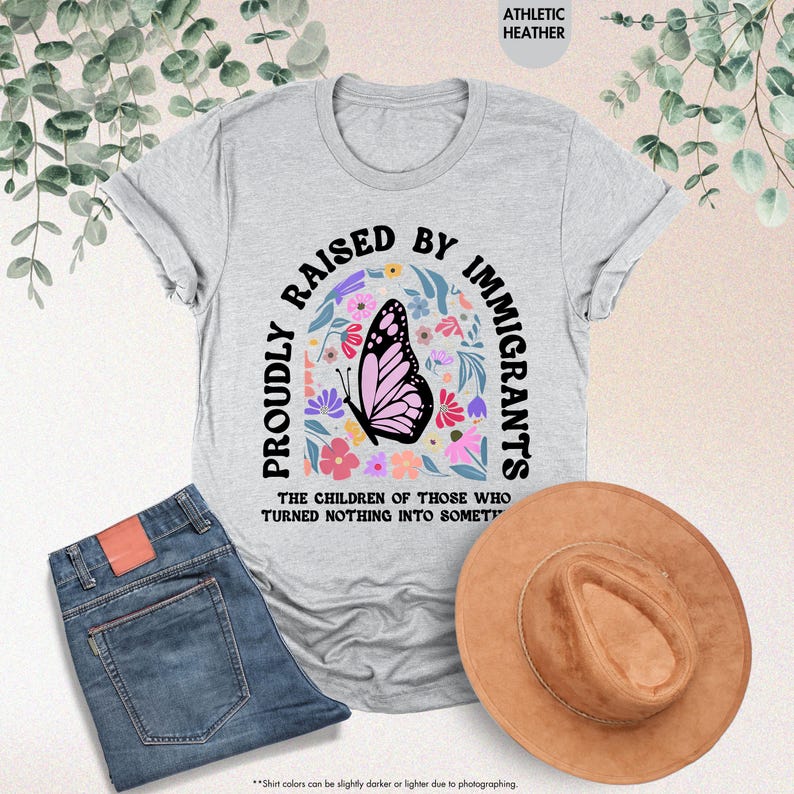 May include: A light gray t-shirt with the text "PROUDLY RAISED BY IMMIGRANTS" and a butterfly graphic. The shirt is paired with blue jeans and a brown hat. The text "THE CHILDREN OF THOSE WHO TURNED NOTHING INTO SOMETHING" is also visible.