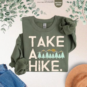K&ouml;nnte beinhalten: Milit&auml;rgr&uuml;nes Sweatshirt mit dem wei&szlig;en Schriftzug "TAKE A HIKE". Das Design umfasst eine Bergkette und B&auml;ume in Gelb- und T&uuml;rkist&ouml;nen. Das Sweatshirt ist gefaltet, wobei die &Auml;rmel sichtbar sind. Der Text "colors can be slightly darker or lighter due to photographing" ist ebenfalls sichtbar.