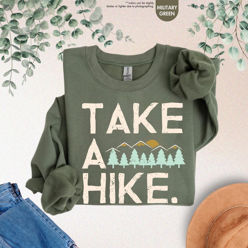K&ouml;nnte beinhalten: Milit&auml;rgr&uuml;nes Sweatshirt mit dem wei&szlig;en Schriftzug "TAKE A HIKE". Das Design umfasst eine Bergkette und B&auml;ume in Gelb- und T&uuml;rkist&ouml;nen. Das Sweatshirt ist gefaltet, wobei die &Auml;rmel sichtbar sind. Der Text "colors can be slightly darker or lighter due to photographing" ist ebenfalls sichtbar.