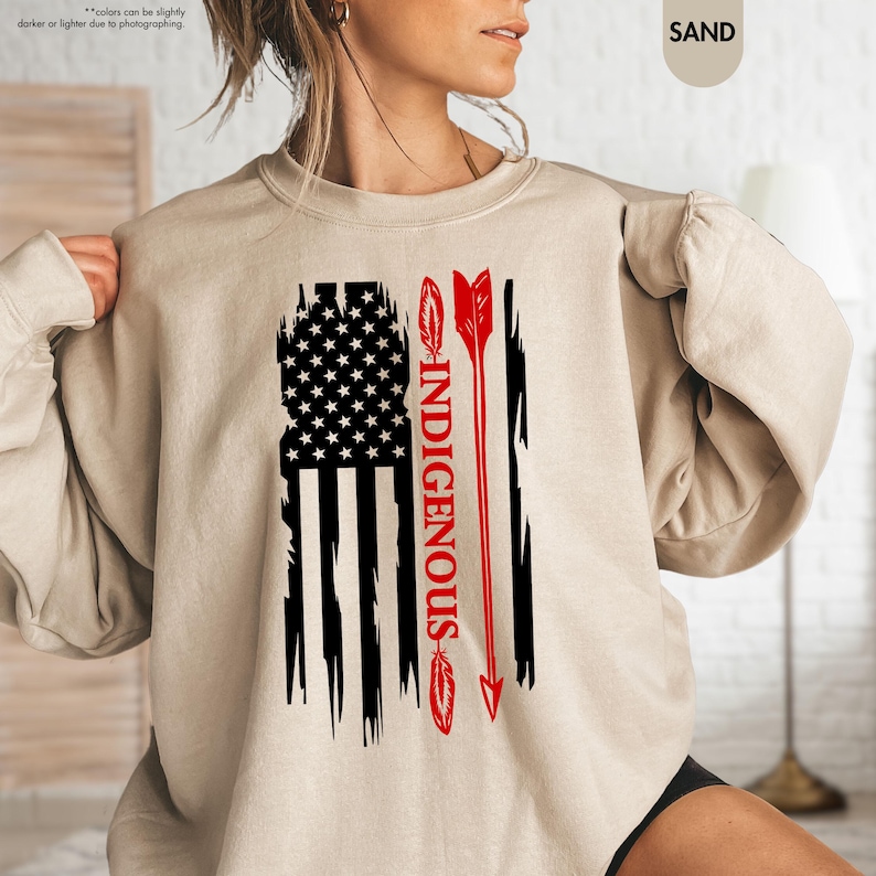 May include: A beige sweatshirt with a distressed American flag design and the word "INDIGENOUS" printed vertically down the center. A red arrow with feathers is on either side of the word.