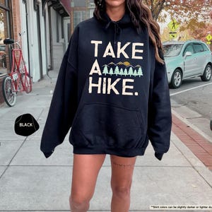 K&ouml;nnte beinhalten: Schwarzer Kapuzenpullover mit dem Text "TAKE A HIKE." in Wei&szlig;, mit einem Berg- und Baum-Motiv. Der Hoodie hat eine Vordertasche und eine Kapuze mit Kordelzug. Das Wort "BLACK" befindet sich in einem schwarzen Kreis unten links.