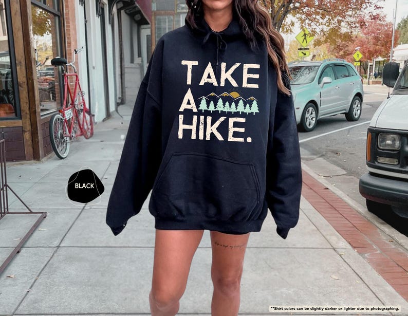 K&ouml;nnte beinhalten: Schwarzer Kapuzenpullover mit dem Text "TAKE A HIKE." in Wei&szlig;, mit einem Berg- und Baum-Motiv. Der Hoodie hat eine Vordertasche und eine Kapuze mit Kordelzug. Das Wort "BLACK" befindet sich in einem schwarzen Kreis unten links.