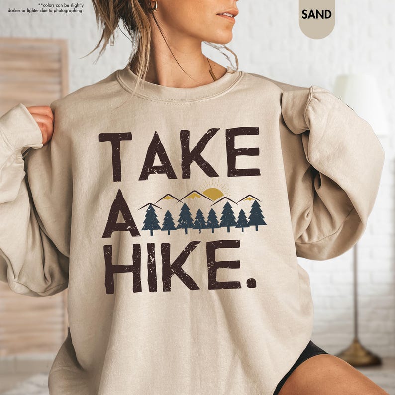 K&ouml;nnte beinhalten: Sandfarbenes Sweatshirt mit dem Text "TAKE A HIKE." in dunkelbraunen Buchstaben. Das Design umfasst eine Bergkette, Sonne und Kiefern. Das Sweatshirt ist ein lang&auml;rmeliger Rundhalsausschnitt.