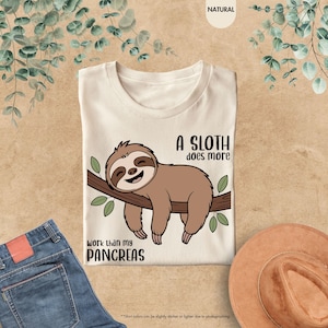 K&ouml;nnte beinhalten: Cremefarbenes T-Shirt mit einem Cartoon-Faultier auf einem Ast. Der Text lautet: "Ein Faultier arbeitet mehr als meine Bauchspeicheldr&uuml;se." Das Design zeigt ein l&auml;chelndes Faultier in Braun- und Beiget&ouml;nen.