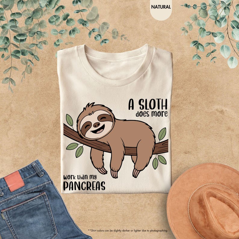 K&ouml;nnte beinhalten: Cremefarbenes T-Shirt mit einem Cartoon-Faultier auf einem Ast. Der Text lautet: "Ein Faultier arbeitet mehr als meine Bauchspeicheldr&uuml;se." Das Design zeigt ein l&auml;chelndes Faultier in Braun- und Beiget&ouml;nen.