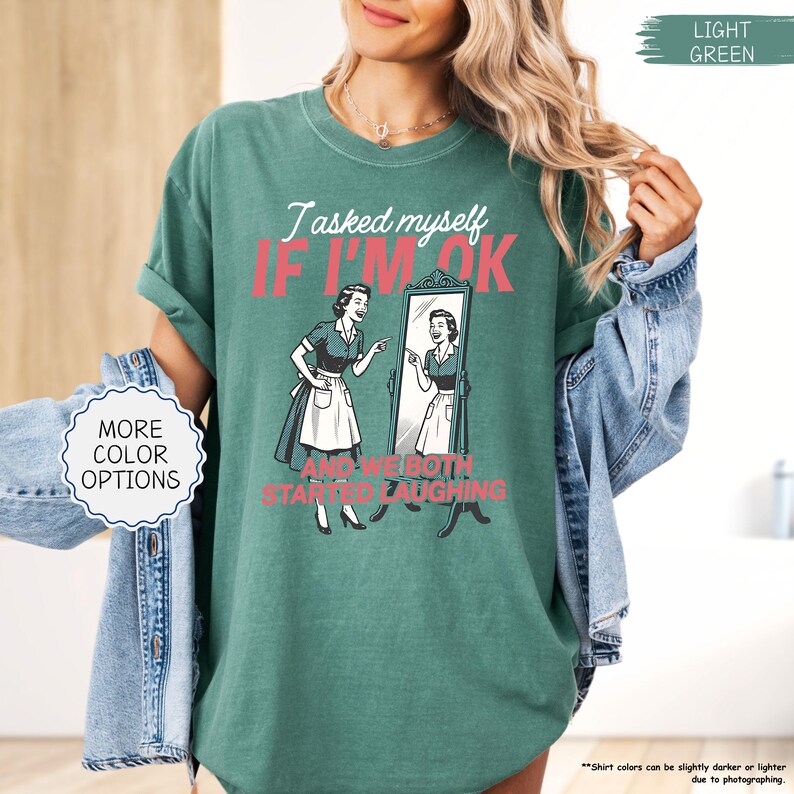 May include: Light green t-shirt with the text "I asked myself if I'm ok and we both started laughing" in pink and black. The graphic shows a vintage illustration of a woman looking in a mirror. The shirt has a relaxed fit.