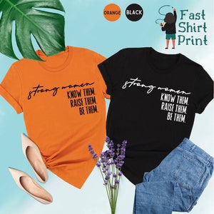 K&ouml;nnte beinhalten: Zwei T-Shirts mit dem Text "strong women know them. raise them. be them." bedruckt. Ein Shirt ist orange und das andere ist schwarz.
