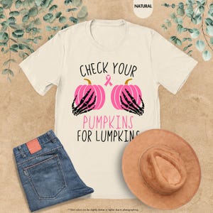 以下が含まれることがあります： 「CHECK YOUR PUMPKINS FOR LUMPKINS」の文字が入ったクリーム色のTシャツ。デザインは、黒い骸骨の手とピンクのリボンが付いた2つのピンク色のカボチャが特徴です。青いジーンズと茶色の帽子も画像にあります。