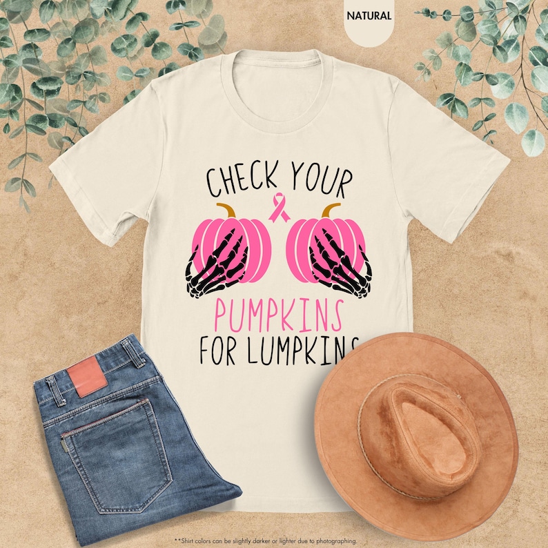 以下が含まれることがあります： 「CHECK YOUR PUMPKINS FOR LUMPKINS」の文字が入ったクリーム色のTシャツ。デザインは、黒い骸骨の手とピンクのリボンが付いた2つのピンク色のカボチャが特徴です。青いジーンズと茶色の帽子も画像にあります。