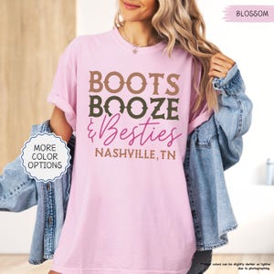 Pu&ograve; includere: T-shirt rosa chiaro con la scritta "BOOTS BOOZE & Besties" in marrone, verde e rosa. Il testo "NASHVILLE, TN" &egrave; sotto. La maglietta ha un girocollo e maniche corte. La parola "BLOSSOM" &egrave; in alto a destra.