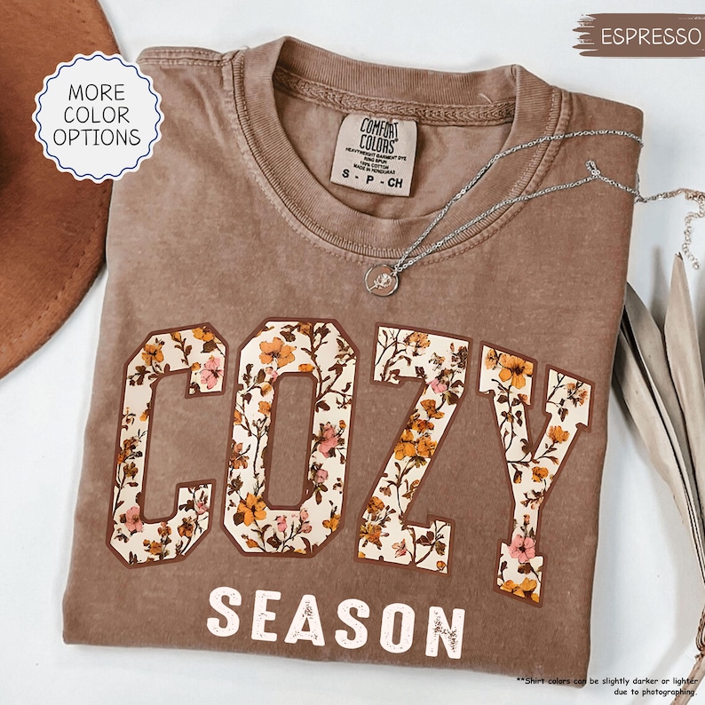 May include: A brown t-shirt with the word "COZY" in large, floral letters and "SEASON" underneath. The shirt has a crew neck and short sleeves. A silver necklace with a small pendant is draped across the shirt. The shirt is labelled "Espresso".