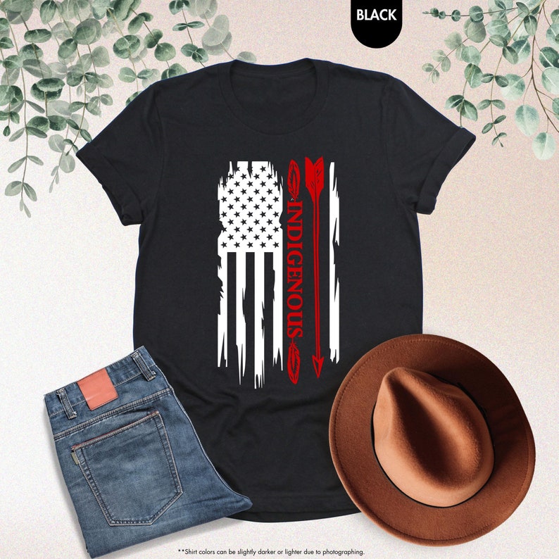 May include: Black t-shirt with a distressed American flag design and the word "INDIGENOUS" written vertically down the right side of the flag. A red arrow with a feather is next to the flag.