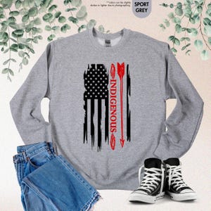 May include: A grey sweatshirt with a distressed American flag design and the word "INDIGENOUS" printed vertically in red. An arrow is shown in red, pointing upwards.