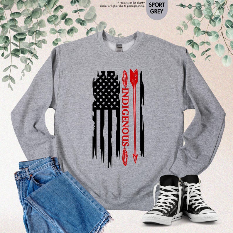 May include: A grey sweatshirt with a distressed American flag design and the word "INDIGENOUS" printed vertically in red. An arrow is shown in red, pointing upwards.