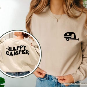 May include: Sand-colored sweatshirt with a black camper graphic on the front and the words "HAPPY CAMPER" on the back. The camper graphic includes a heart. The sweatshirt is a neutral color and suitable for casual wear.