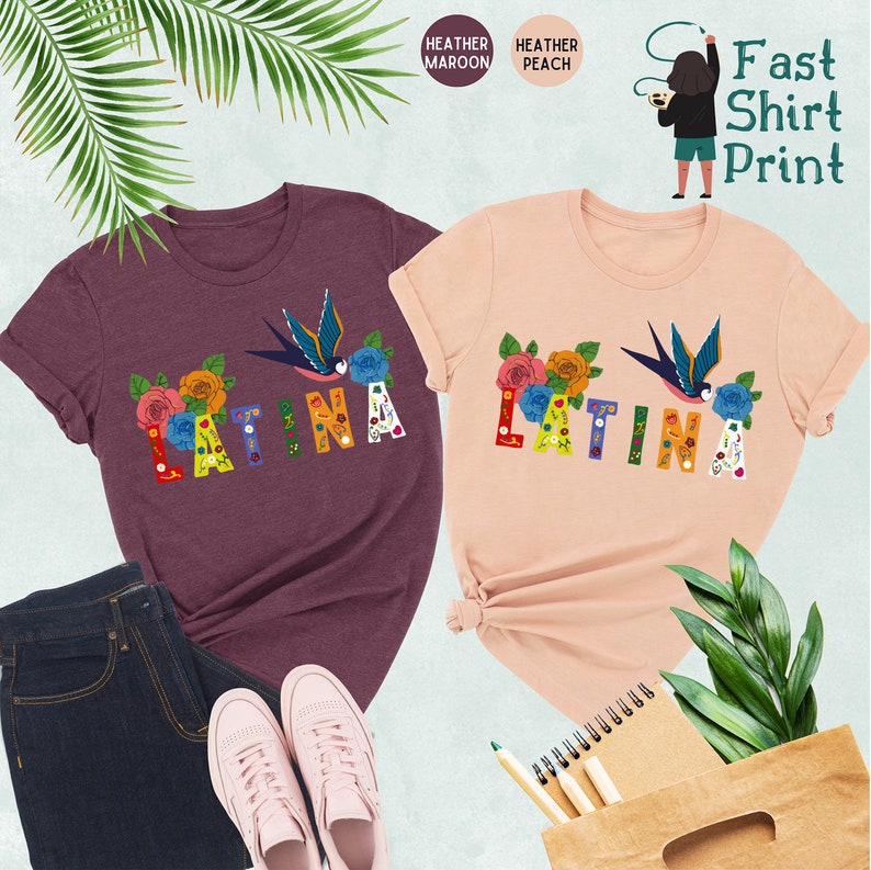 May include: Two t-shirts with the word "LATINA" in colorful letters decorated with flowers and a bird. The shirts are available in heather maroon and heather peach.