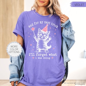 May include: A violet short-sleeved t-shirt featuring a white graphic of a kitten wearing a party hat and holding a star wand. The text reads, and for my next trick i'll forget what i was doing, surrounded by small stars and sparkles.