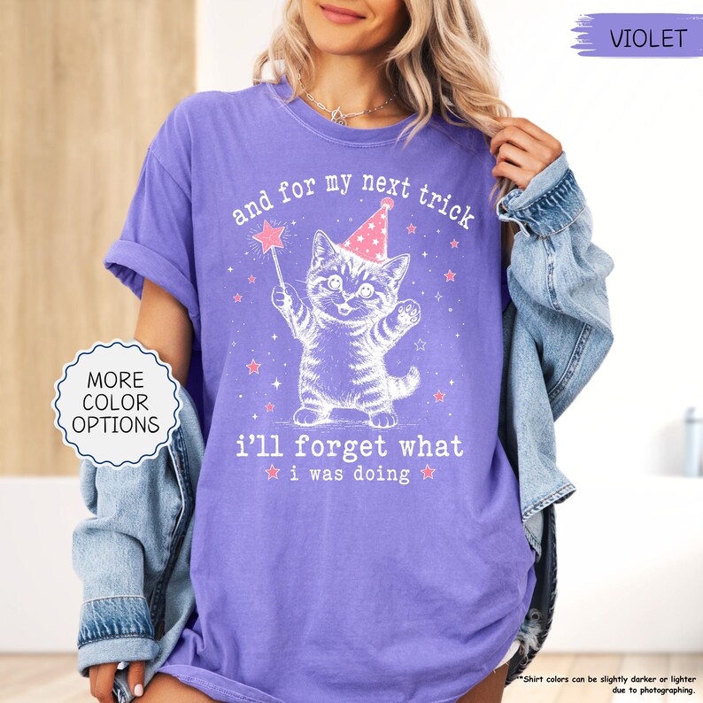 May include: A violet short-sleeved t-shirt featuring a white graphic of a kitten wearing a party hat and holding a star wand. The text reads, and for my next trick i'll forget what i was doing, surrounded by small stars and sparkles.