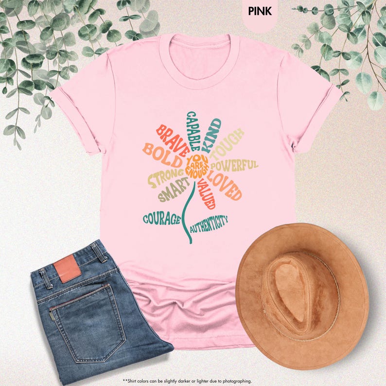 Pode incluir: Uma camiseta rosa com um design floral com palavras como "Corajoso", "Forte" e "Amado". A camiseta &eacute; combinada com jeans azuis e um chap&eacute;u marrom. O texto "PINK" est&aacute; no topo.