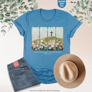May include: A blue t-shirt with a floral design and a cross on a hill. The text "He is not here. He has risen. Matthew 28:6" is printed on the shirt.