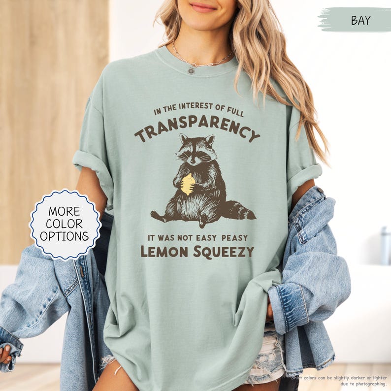 May include: A light green t-shirt with a raccoon graphic holding a lemon. The text on the shirt reads "IN THE INTEREST OF FULL TRANSPARENCY, IT WAS NOT EASY PEASY LEMON SQUEEZY". The shirt also has a "MORE COLOR OPTIONS" button.