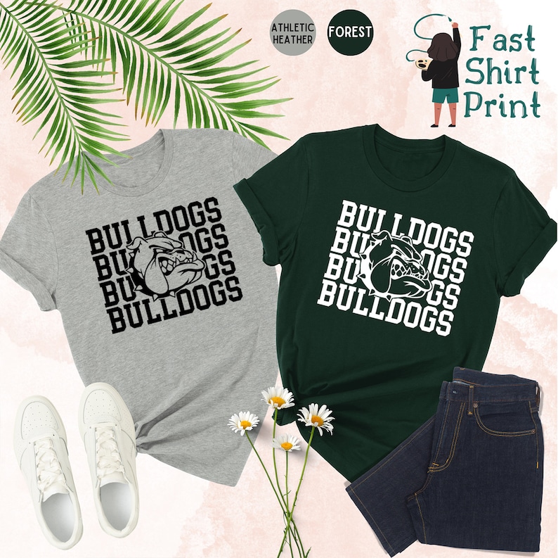 May include: Two t-shirts with the word "Bulldogs" repeated in a stacked pattern. The shirts are in athletic heather grey and forest green. The word "Bulldogs" is in black on the grey shirt and white on the green shirt. A bulldog mascot is featured in the center of each design.