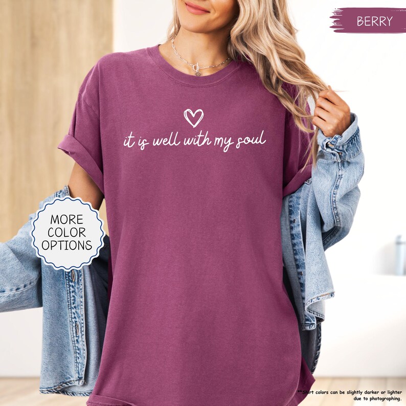K&ouml;nnte beinhalten: Beerenfarbenes T-Shirt mit dem Schriftzug "it is well with my soul" und einem Herz-Grafik. Das Shirt hat hochgekrempelte &Auml;rmel und wird mit einer Jeansjacke getragen. Ein Knopf auf der linken Seite zeigt "MORE COLOR OPTIONS".