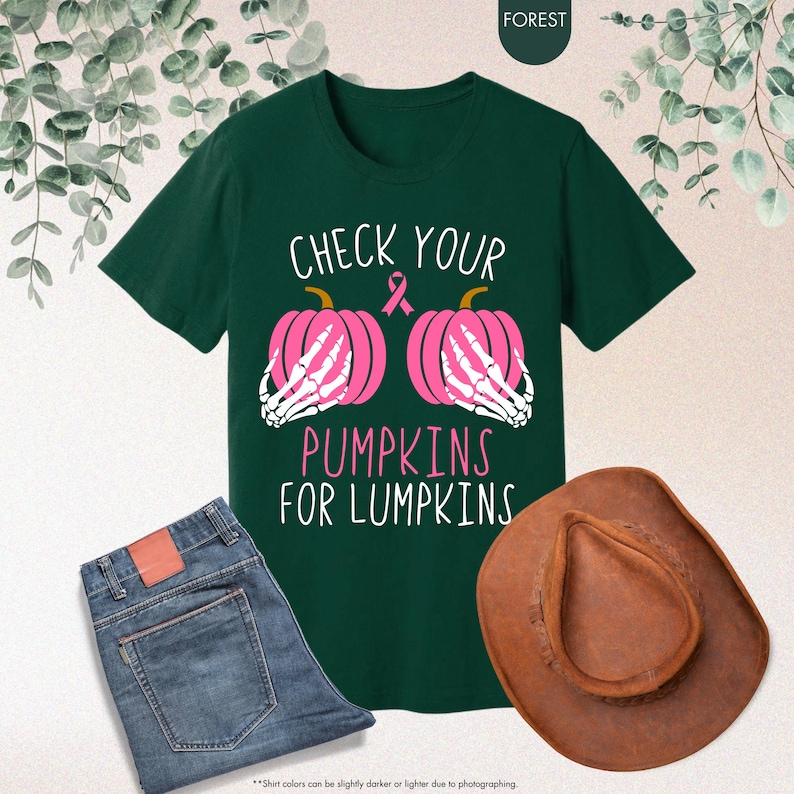 以下が含まれることがあります： 「CHECK YOUR PUMPKINS FOR LUMPKINS」の文字と、骸骨の手を持つピンク色のカボチャ2つのグラフィックがプリントされた濃い緑色のTシャツ。画像には、青いジーンズと茶色のカウボーイハットも含まれています。