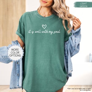K&ouml;nnte beinhalten: Ein hellgr&uuml;nes T-Shirt mit dem Text "it is well with my soul" in Schreibschrift und einem Herzsymbol. Das T-Shirt hat hochgekrempelte &Auml;rmel und ist Teil einer Bekleidungskollektion. Die Worte "MORE COLOR OPTIONS" sind ebenfalls sichtbar.