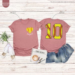 K&ouml;nnte beinhalten: Zwei T-Shirts in der Farbe Heide-Mauve mit einem Softball-Design. Das vordere T-Shirt hat einen herzf&ouml;rmigen Softball mit dem Namen "Margaret" darunter. Das hintere T-Shirt hat eine gro&szlig;e Nummer "10" mit einem Softball-Design.