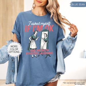 May include: Blue jean colored t-shirt with the text "I asked myself if I'm ok and we both started laughing" and a retro illustration of a woman looking in a mirror. The shirt also has the words "More color options" on the left side.
