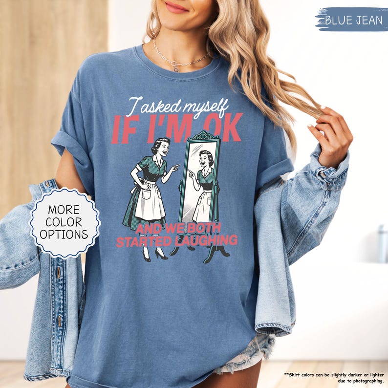 May include: Blue jean colored t-shirt with the text "I asked myself if I'm ok and we both started laughing" and a retro illustration of a woman looking in a mirror. The shirt also has the words "More color options" on the left side.
