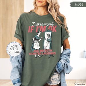 May include: Moss green t-shirt with the text "I asked myself IF I'M OK AND WE BOTH STARTED LAUGHING" and a vintage illustration of a woman looking in a mirror. The shirt also has "MORE COLOR OPTIONS" text.