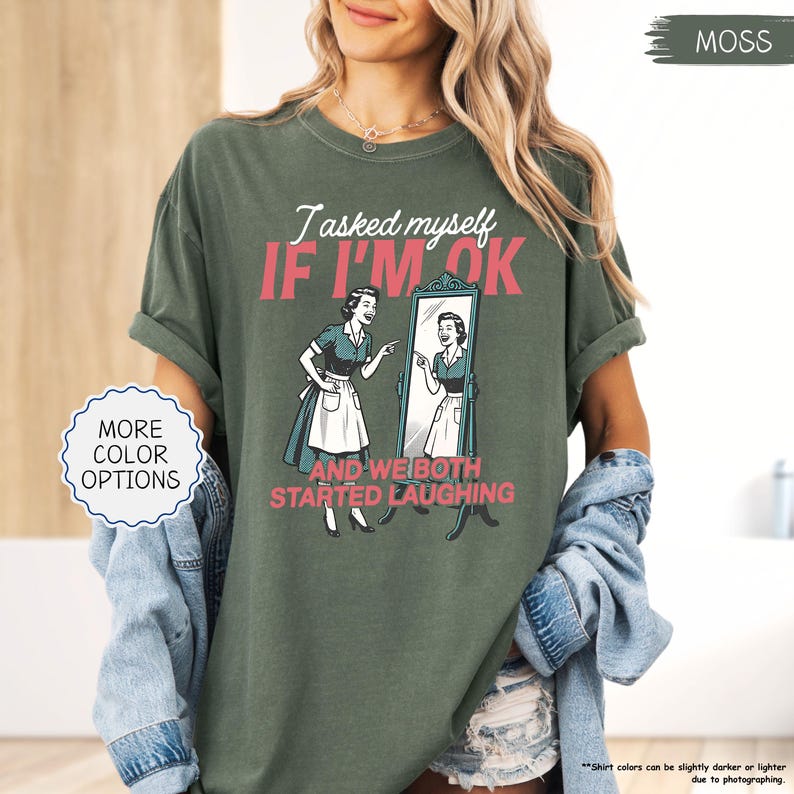 May include: Moss green t-shirt with the text "I asked myself IF I'M OK AND WE BOTH STARTED LAUGHING" and a vintage illustration of a woman looking in a mirror. The shirt also has "MORE COLOR OPTIONS" text.