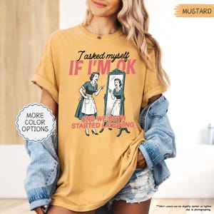 May include: Mustard-colored t-shirt with the text "I asked myself IF I'M OK" and a vintage illustration of a woman looking in a mirror. The shirt also has the text "AND WE BOTH STARTED LAUGHING".