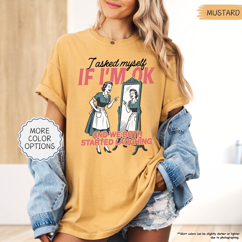 May include: Mustard-colored t-shirt with the text "I asked myself IF I'M OK" and a vintage illustration of a woman looking in a mirror. The shirt also has the text "AND WE BOTH STARTED LAUGHING".