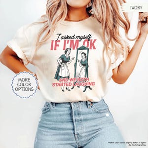 May include: Ivory-colored t-shirt featuring the text "I asked myself IF I'M OK and we both started laughing" with a retro illustration of a woman looking in a mirror. The shirt has a crew neck and short sleeves. More color options are available.