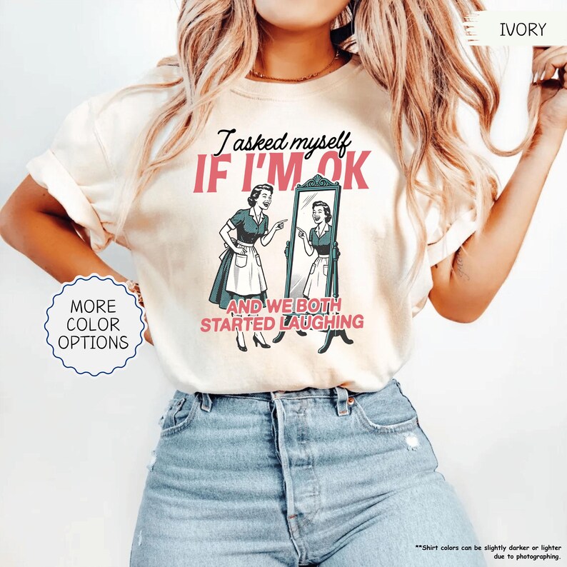 May include: Ivory-colored t-shirt featuring the text "I asked myself IF I'M OK and we both started laughing" with a retro illustration of a woman looking in a mirror. The shirt has a crew neck and short sleeves. More color options are available.