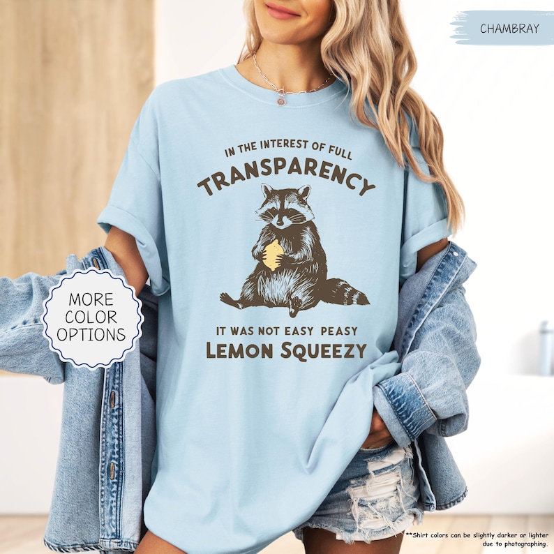 May include: Light blue t-shirt with the text "IN THE INTEREST OF FULL TRANSPARENCY" and "IT WAS NOT EASY PEASY LEMON SQUEEZY". The shirt features a raccoon graphic holding a lemon. The shirt is made of a soft material.