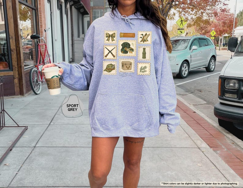 K&ouml;nnte beinhalten: Ein hellgrauer Kapuzenpullover mit K&auml;ngurutasche und Kapuze mit Kordelzug. Auf der Vorderseite des Hoodies befinden sich Briefmarken-Grafiken mit Jagd-Illustrationen. Der Text "SPORT GREY" ist auf der Vorderseite aufgedruckt.
