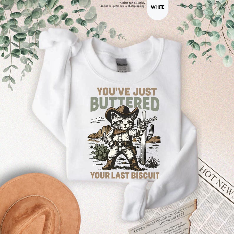 May include: White sweatshirt with a cartoon cat cowboy design, holding a gun. The text reads "YOU'VE JUST BUTTERED YOUR LAST BISCUIT". The graphic includes desert elements like cacti and mountains.