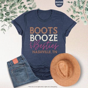 Pu&ograve; includere: Una maglietta blu navy con le parole "BOOTS BOOZE & Besties NASHVILLE, TN" in lettere marroni, bianche e rosa. La maglietta &egrave; abbinata a jeans blu e un cappello da cowboy marrone.