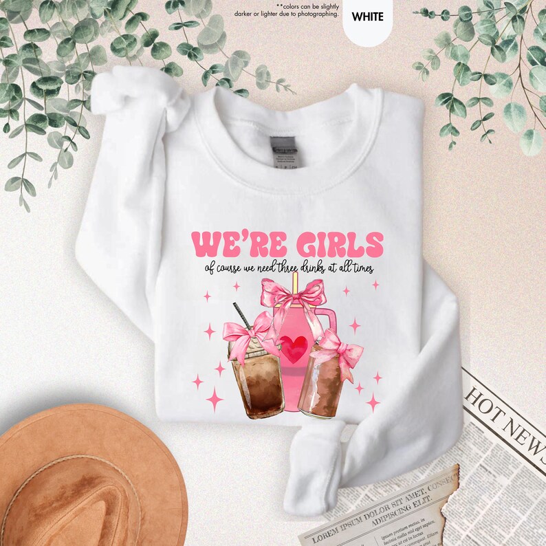 K&ouml;nnte beinhalten: Wei&szlig;es T-Shirt mit einem rosafarbenen Grafikdesign, auf dem "We're Girls of course we need three drinks at all times" steht. Die Grafik zeigt drei rosafarbene Getr&auml;nke mit rosafarbenen Schleifen und Sternen.