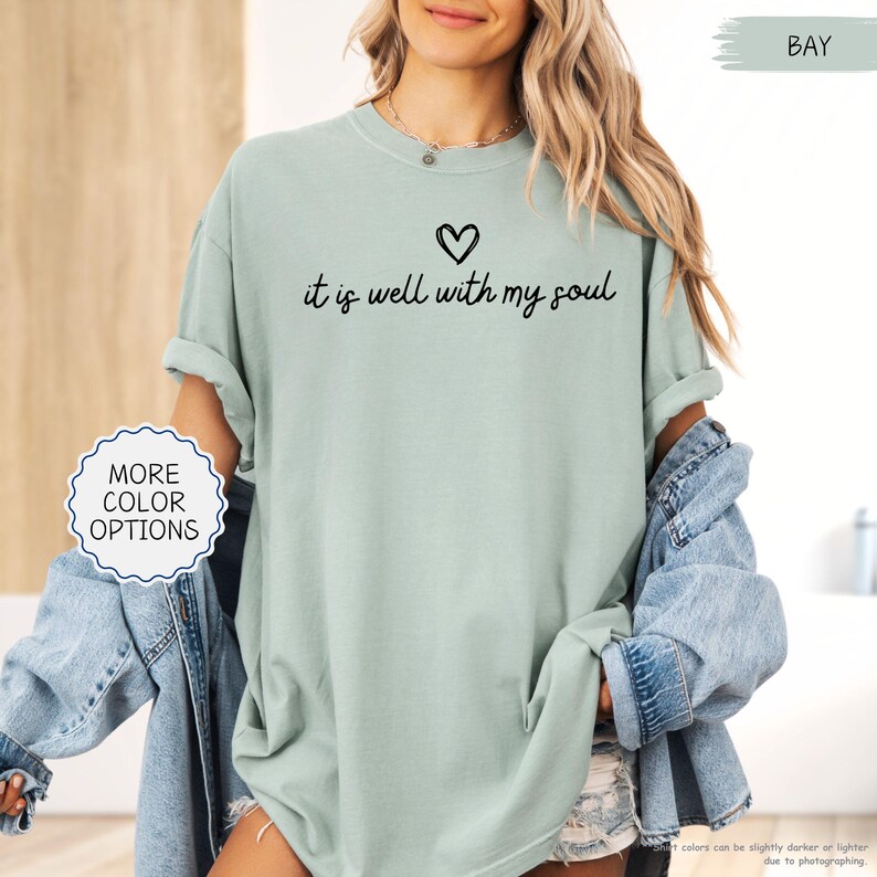 K&ouml;nnte beinhalten: Hellgr&uuml;nes T-Shirt mit dem Schriftzug "it is well with my soul" in schwarzer Schrift, &uuml;ber einem kleinen Herzen. Das Shirt hat hochgekrempelte &Auml;rmel und ist Teil einer Bekleidungskollektion. Eine Jeansjacke ist ebenfalls zu sehen.