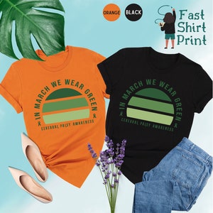 May include: Two t-shirts with a green and white striped graphic that says "In March We Wear Green" and "Cerebral Palsy Awareness". One shirt is orange and the other is black.