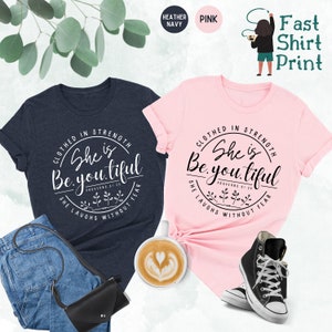 May include: Two t-shirts, one navy and one pink, with the text "Clothed in Strength, She is Beautiful" in a circular design. The navy shirt is paired with jeans and a black purse, while the pink shirt is styled with sneakers.