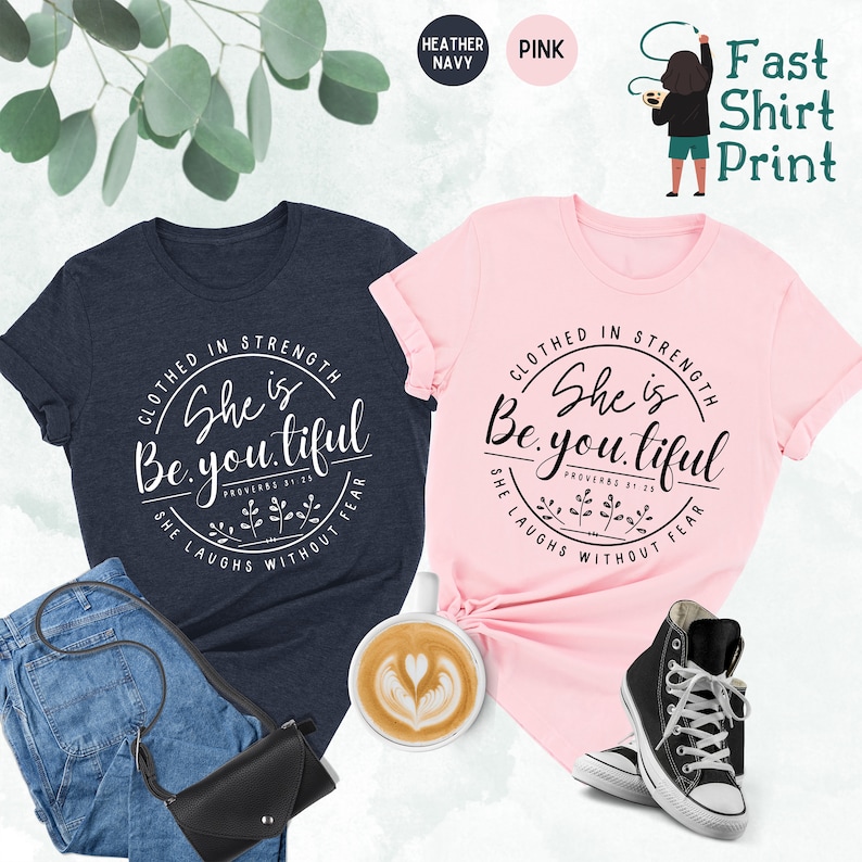 May include: Two t-shirts, one navy and one pink, with the text "Clothed in Strength, She is Beautiful" in a circular design. The navy shirt is paired with jeans and a black purse, while the pink shirt is styled with sneakers.