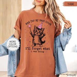 May include: A rust-coloured t-shirt featuring a graphic of a kitten wearing a pink party hat and holding a magic wand. The text reads, "and for my next trick i'll forget what i was doing," surrounded by small pink stars and black sparkles.