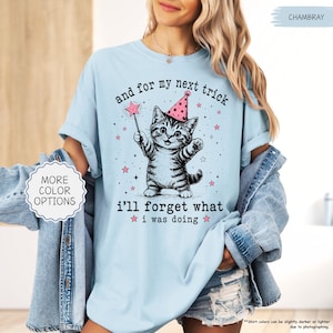 May include: A light blue t-shirt featuring a graphic of a kitten wearing a pink party hat and holding a star-tipped wand. The text reads, "and for my next trick i'll forget what i was doing," surrounded by small pink stars and black sparkles.