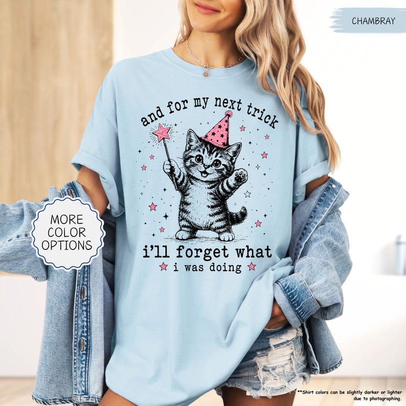 May include: A light blue t-shirt featuring a graphic of a kitten wearing a pink party hat and holding a star-tipped wand. The text reads, "and for my next trick i'll forget what i was doing," surrounded by small pink stars and black sparkles.